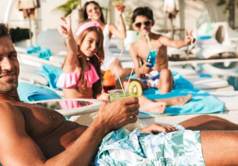 Eine Familie ist im Urlaub am Pool. Zu sehen sind im Hintergrund eine Frau, in der mitte 2 Kinder und vorne ein Mann, der sich bisschen Abstand gönnt