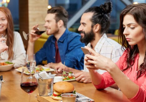 Ein paar Freunde sitzen am Tisch und die vorderste Frau schaut abwesend auf ihr Handy