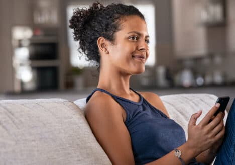 eine Frau sitzt auf dem Sofa, ihr Handy in der Hand. Sie schaut nachdenklich aber zufrieden aus